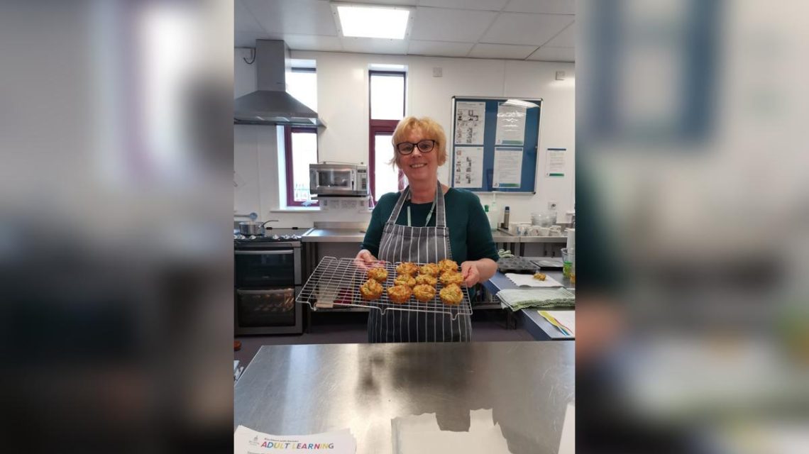 A woman holding a tray of muffins, smiling in a kitchen.